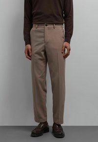 Pantalon beige à coupe ajustée, confectionné dans un tissu lisse, avec des poches latérales et une fermeture à bouton unique, assorti à des chaussures foncées.