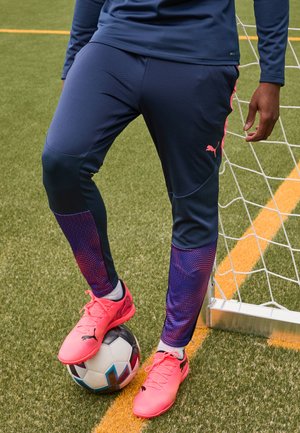Marineblauwe sportbroek met roze accenten, textuurmateriaal en een aansluitend ontwerp; gecombineerd met felroze voetbalschoenen op een zwart-witte voetbal.