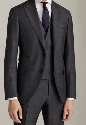 Homme portant un costume trois pièces à carreaux gris foncé sur mesure avec une chemise blanche et une cravate bleu marine foncé sur un fond clair uni.