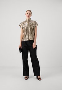 Top dorato con paillettes e colletto alto, abbinato a pantaloni neri con righe sottili. L'outfit è completato da una pochette nera e sandali con tacco a blocco.