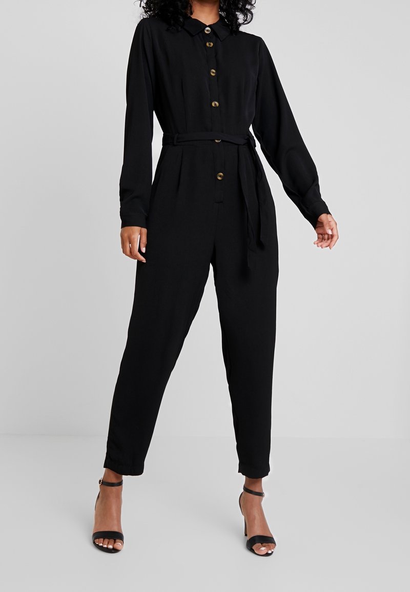 Femme portant une combinaison noire à manches longues avec boutons sur le devant, ceinture à la taille, et escarpins noirs à bride cheville, debout devant un fond uni.