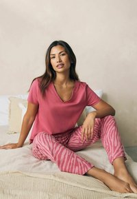 Rosa V-Ausschnitt-T-Shirt kombiniert mit rosa gestreiften Hosen im Zebramuster. Weicher Stoff, lockere Passform, auf einer strukturierten beigen Decke sitzend.