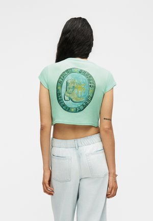 Γυναίκα με μακριά σκούρα μαλλιά φοράει κοντό μωβ πουκάμισο σε χρώμα μέντας με γραφικό καουμπόικης μπότες και ήλιο στην πλάτη και ανοιχτό μπλε παντελόνι με ψηλή μέση.