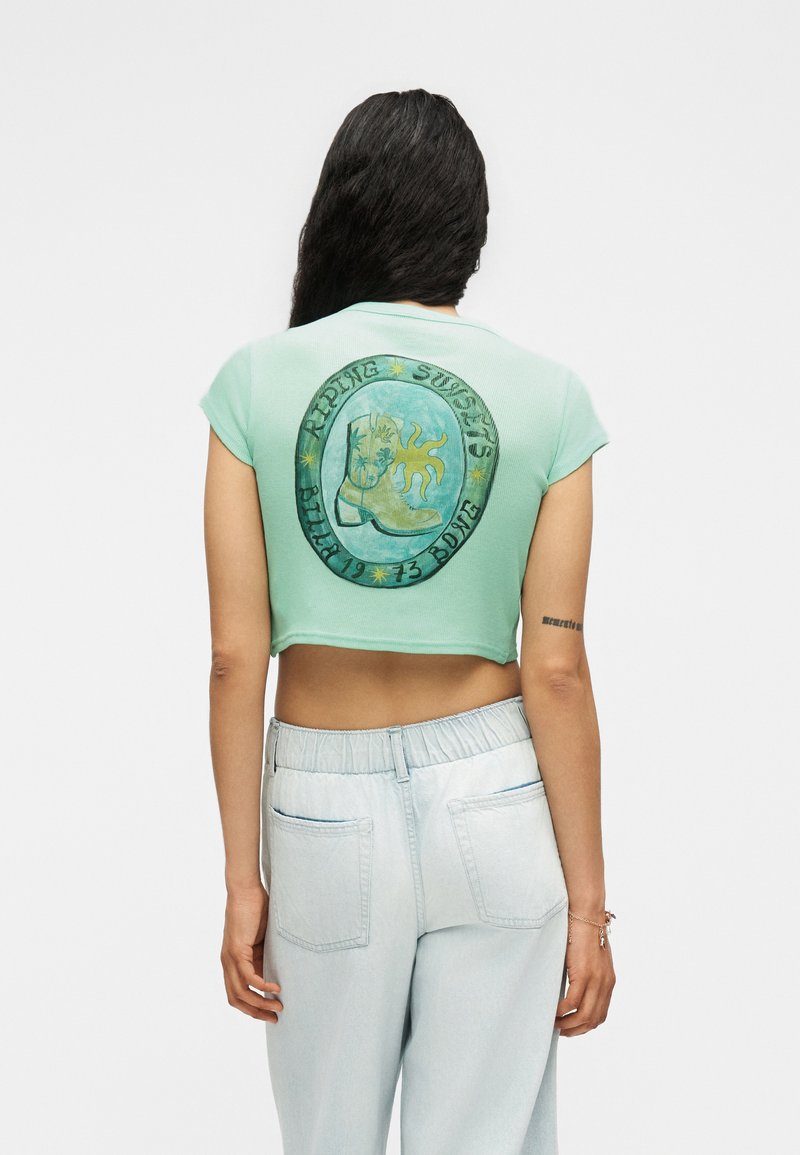 Γυναίκα με μακριά σκούρα μαλλιά φοράει κοντό μωβ πουκάμισο σε χρώμα μέντας με γραφικό καουμπόικης μπότες και ήλιο στην πλάτη και ανοιχτό μπλε παντελόνι με ψηλή μέση.