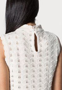 Sweter w kolorze kremowym, z wzorem w diamenty, falbankowymi brzegami i zapięciem na guziki z tyłu, z łezkowatym otworem.