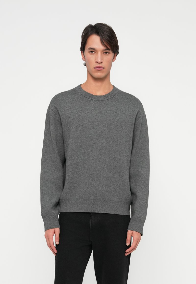 Grå strikkegenser med rund hals, lange ermer og ribbestrikket kant. Brukt over mørke jeans, har en glatt tekstur og avslappet passform.