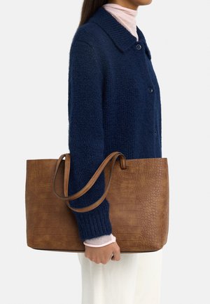 Kobieta w granatowym swetrze i białych spodniach trzyma przy boku dużą brązową torebkę na ramię z teksturowanej skóry.