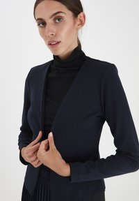 Námorně modrý blazer s dlouhým rukávem a přiléhavým střihem, vyrobený z měkkého materiálu, s hlubokým výstřihem do V a minimalistickým designem. Texturovaný černý rolák pod ním.