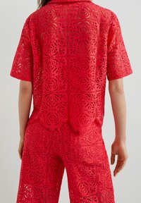 Vue arrière d'une personne portant un haut à manches courtes en dentelle rouge et un pantalon assorti avec des motifs découpés floraux et géométriques.