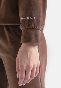 Brązowy, miękki dres z marszczonymi mankietami. Na lewym mankiecie wyhaftowany tekst "Łatwe do pokochania!". Gładka faktura i dopasowany krój.