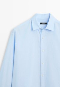 Světle modrá košile s dlouhým rukávem vyrobená z hladké tkaniny. Obsahuje klasický límeček, zapínání na knoflíky a jednu kapsu na hrudi. Jednoduchá textura.