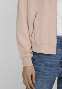 Light pink bomber jacket with ribbed cuffs and hem, featuring a side zip pocket and smooth fabric texture, paired with blue denim jeans.
