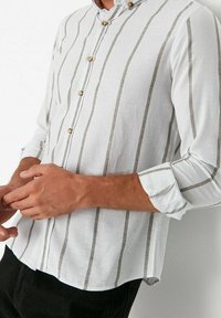 Valkoinen napillinen paita, jossa pystysuorat harmaat raidat, ruskeat napit ja käärittyjä hihoja, valmistettu kevyestä kankaasta.