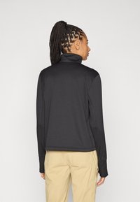 Μαύρο μπουφάν με φερμουάρ και ψηλό γιακά, υφασμάτινη υφή, μακριά μανίκια και άνετη εφαρμογή, συνδυασμένο με καφέ παντελόνια.