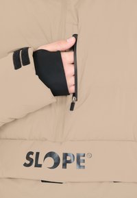 Beige pufferjacka med en dragkedjeficka, svart innerfoder och en framträdande svart "SLOPE"-logotyp vid fållen. Texturerad material.