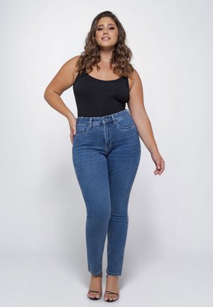 Krullend bruinharige vrouw in een zwart tanktop, blauwe high-waist jeans en zwarte sandalen met bandjes staat tegen een witte achtergrond.