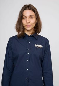 Tengerészkék gombos ing hosszú ujjakkal, mellkasi zseb fehér csipkével és kis barna szalag díszítéssel. Simább anyagú textúra.