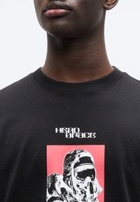 Μαύρο βαμβακερό μπλουζάκι με έντονο γραφικό εκτύπωμα ενός αστροναύτη σε λευκό πάνω σε ροζ φόντο, με το κείμενο "HEAD SPACE" από πάνω.