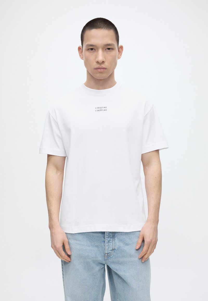 Νέος με κοντά μαλλιά που φοράει ένα λευκό T-shirt με την επιγραφή «LIBERTINE» και ανοιχτόχρωμο τζιν, στέκεται μπροστά σε ένα απλό λευκό φόντο.