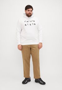 Biała bluza z kapturem z logo "CALVIN KLEIN", beżowe spodnie i czarne sneakersy. Bluza ma regulowany kaptur i ściągacze przy mankietach.