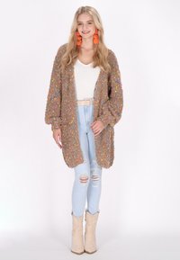 Chunky, texturerad cardigan i beige med färgglada inslag, öppen framsida och ballongärmar; kombinerad med ljusblå slitna jeans och beiga stövlar.