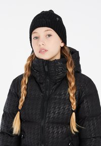Giubbotto imbottito nero con motivo pied de poule e colletto alto, abbinato a un berretto nero a coste con dettaglio del logo. Due tasche con cerniera.