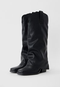 Paire de bottes hautes en cuir noir avec talons carrés bas et languettes pour enfiler, placées côte à côte sur un fond blanc.