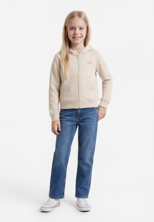 Fată zâmbitoare în picioare cu o mână pe șold, purtând hanorac bej cu fermoar, blugi albaștri și adidași albi pe fundal alb.