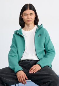 Turquoise Nike zip-up hoodie with a white interior lining and black accents, worn over a white t-shirt, paired with dark gray pants.