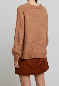 Pull-over marron clair à coupe décontractée, avec poignets et ourlet côtelés. Associé à une jupe en velours côtelé rouille foncé, texturée de lignes verticales.