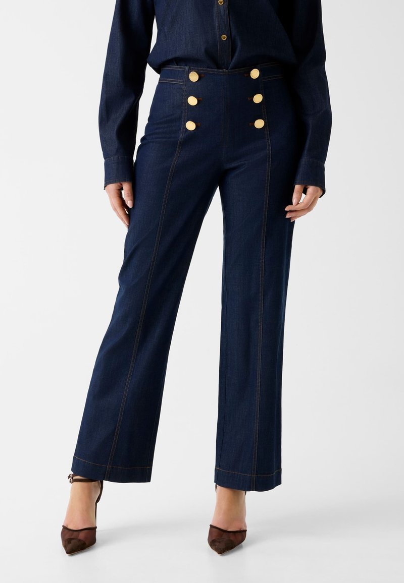 Frau, die dunkelblaue, hoch taillierte, weit geschnittene Hose mit sechs goldenen Knöpfen und dunkelbraunen spitzen Absätzen trägt, steht vor einem einfarbigen Hintergrund.