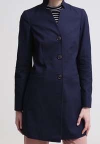 Abrigo entallado de color azul marino con diseño sin cuello, cierre de botones frontal y dos bolsillos laterales. Tejido suave y silueta estructurada.