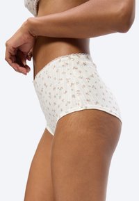 Femme portant une culotte taille haute blanche à petit motif floral rose et bordure en dentelle, montrant un profil latéral de la taille jusqu'à mi-cuisse.
