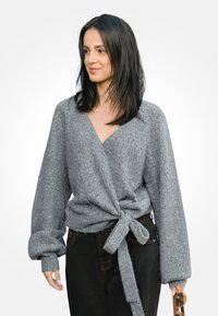 Suéter gris envolvente con cuello en V, tejido texturizado y cintura atada. Combinado con jeans oscuros. El modelo sostiene unas gafas de sol en una mano.