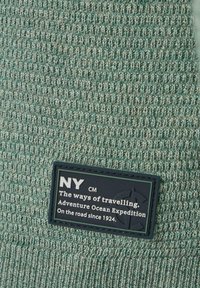 Gestrickter Pullover in hellem Petrol mit einem strukturierten Muster. Verfügt über ein schwarzes Stoffetikett mit weißem Text und einem Emblem, das die Marke und Details angibt.