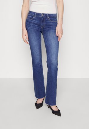 Jeans de denim azul de campana con una cintura de altura media, que cuentan con una cremallera, bolsillos delanteros y un dobladillo ligeramente deshilachado. Combinados con tacones negros.