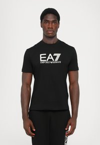 Sort t-shirt lavet af blødt bomuld, med et fremtrædende hvidt EA7 Emporio Armani-logo på forsiden. Kortærmet og rundhalset design.
