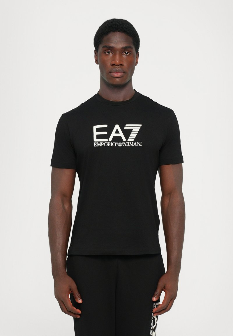 Sort t-shirt lavet af blødt bomuld, med et fremtrædende hvidt EA7 Emporio Armani-logo på forsiden. Kortærmet og rundhalset design.