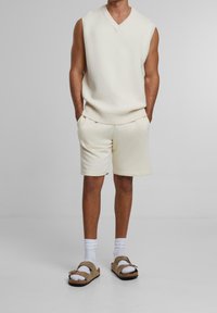 Pull sans manches crème avec col en V ; short assorti crème ; sandales marron à double lanière et chaussettes blanches. Texture lisse sur toute la surface.