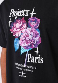 Camiseta negra con un gráfico floral colorido que incluye peonías e hidrangeas, junto con elementos de texto en blanco y púrpura.
