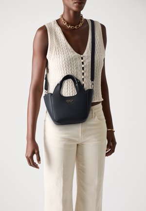 Borsa a tracolla in pelle nera con texture, dotata di doppi manici e una tracolla staccabile. Dettaglio del logo oro sul frontale. Indossata con un top in maglia beige.