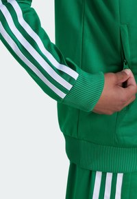 Green athletic jacket with white stripes along sleeves. Features a zippered pocket, ribbed cuffs, and smooth texture.