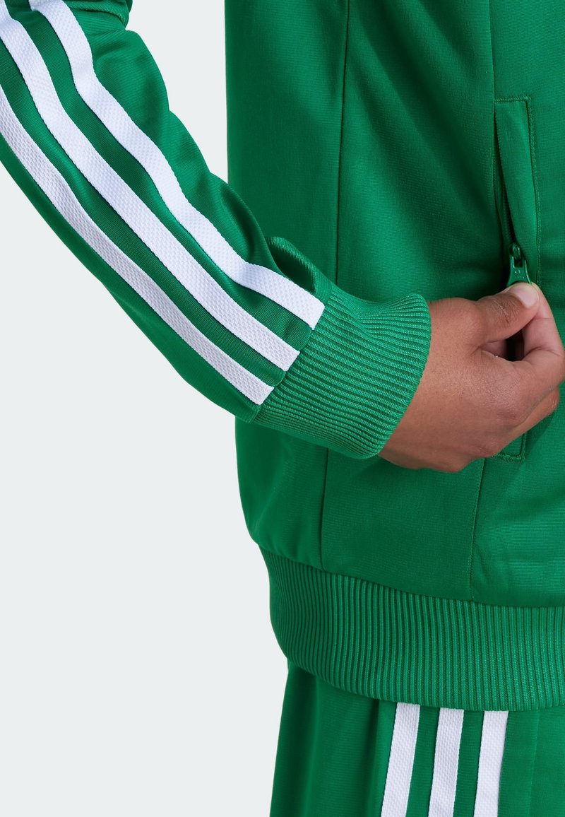 Green athletic jacket with white stripes along sleeves. Features a zippered pocket, ribbed cuffs, and smooth texture.