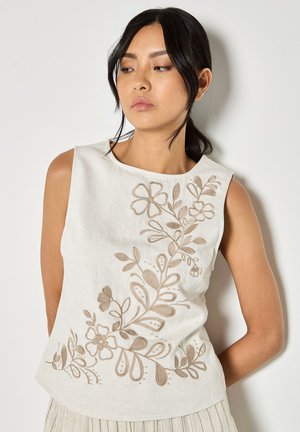 Femme aux cheveux foncés attachés en queue de cheval basse, portant un haut blanc sans manches avec une broderie florale beige, debout devant un mur clair uni.