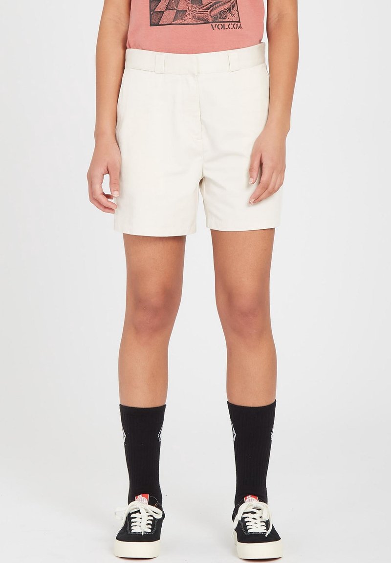 Personne portant un short blanc sur mesure, des chaussettes noires, des baskets noires et blanches, et un t-shirt graphique rose, visible de la taille vers le bas.