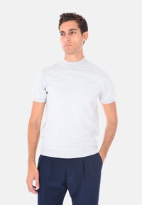 Uomo che indossa un maglione corto grigio chiaro con maniche corte e pantaloni plissettati blu scuro, in piedi con una mano in tasca su uno sfondo bianco.
