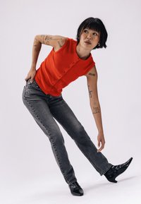 Haut sans manches en tricot côtelé rouge avec des détails de boutons, associé à un jean en denim gris. L'individu porte des bottines noires à bout pointu.