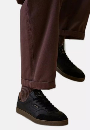 Gambe con pantaloni marroni e calzini marroni a coste con sneakers nere Faguo con suola in gomma su sfondo chiaro.