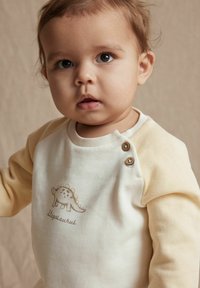 Sweat-shirt pour bébé crème et jaune avec imprimé dinosaure, boutons à l'épaule, texture douce, col rond et manches longues.
