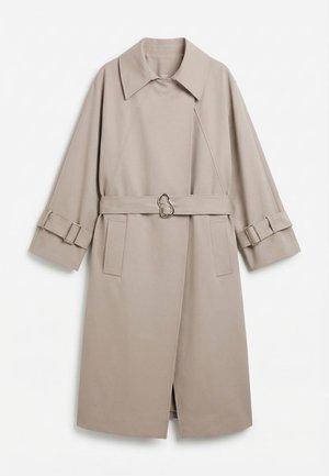 Μπεζ μακρύ παλτό τύπου τρανς με φαρδιά γιακά, ζώνη στη μέση με μοναδικό μεταλλικό αγκράφα και ταιριαστά μανίκια με μεταλλικά στοιχεία.